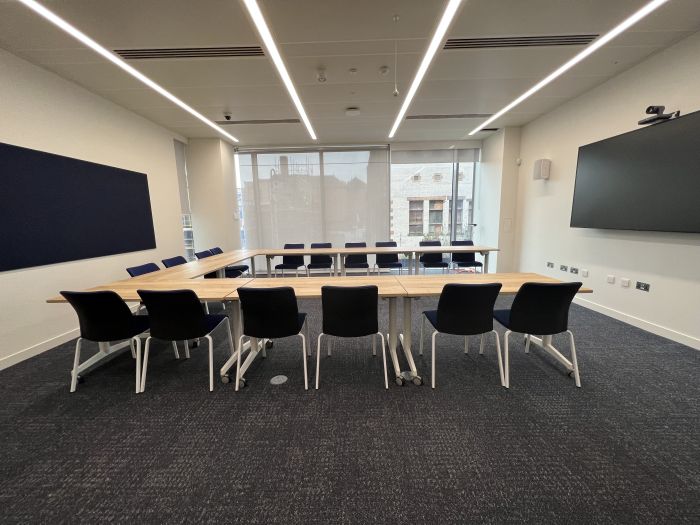 Flat-floored meeting room with tables and chairs in a horseshoe layout, wall-mounted video monitor, videoconferencing camera, speakers and microphone.