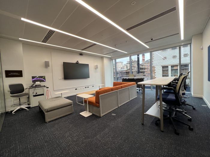 Flat-floored meeting room with high tables and chairs and sofa arranged in parallel L-shape layout, wall-mounted video monitor, videoconferencing camera, microphone and speakers, lectern, coffee table, various side tables, and low padded bench..