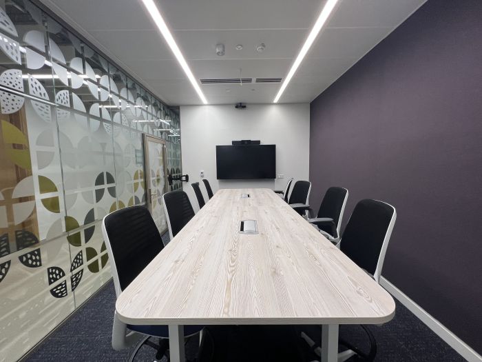 Flat-floored meeting room with high boardroom table and chairs, and wall mounted video monitor.