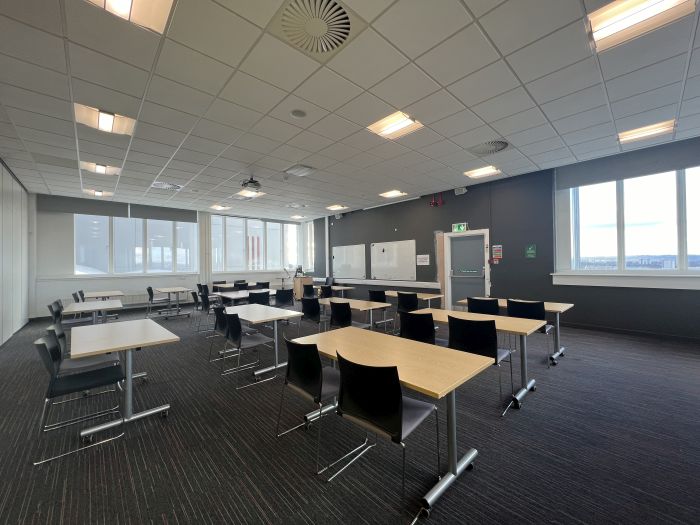 Flat floored teaching room with tables and chairs in rows and groups, whiteboards, visualiser, lectern, projector, lecturer's chair, and PC.