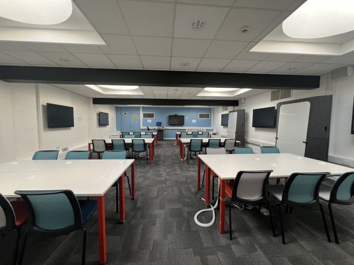 Flat floored teaching room with rows of tables and chairs, moveable whiteboards, video monitors, lectern, and PC.