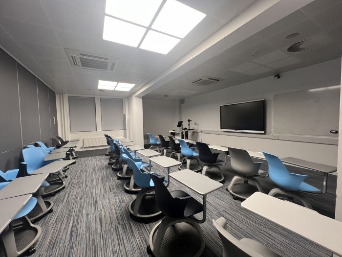 Flat floored teaching room with rows of tablet chairs, smart screen, whiteboard, visualiser, lectern, and PC.