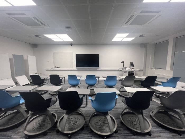 Flat floored teaching room with rows of tablet chairs, smart screen, wall mounted whiteboard, moveable whiteboards, visualiser, lectern, and PC.