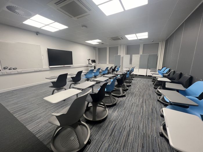 Flat floored teaching room with rows of tablet chairs, smart screen, wall mounted whiteboard, moveable whiteboards, visualiser, lectern, and PC.