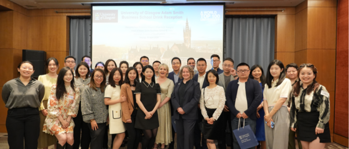 Head of School Eleanor Shaw and Dr Xiang Li with a group of alumni in Beijing, China. They stand before a large powerpoint presentation showing the Gilmorehill campus tower.