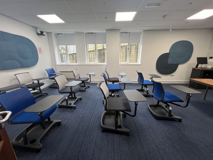 Flat floored teaching room with tablet chairs, acoustic wall panelling, lecturer's desk, lectern, and PC.