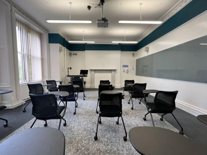 Flat floored teaching room with tablet chairs, glassboard, visualiser, PC, lectern, and projector.