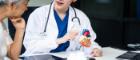 A young female nurse works at a desk with a grey-haired Asian mature woman, discussing heart-related conditions like heart disease, heart attack, and hypertension.