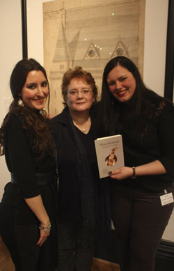 three women (the researchers) stand in the museum in front of an architectural drawing. One holds a copy of the project publication 