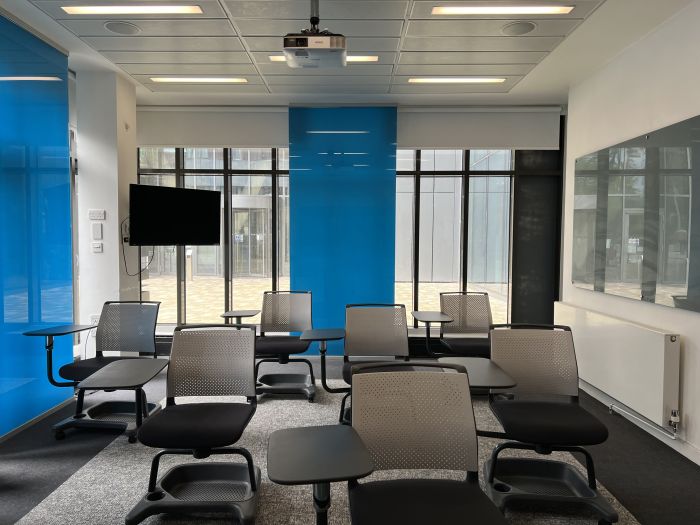 Flat floored teaching room with tablet chairs, large monitor, projector, and glassboards.