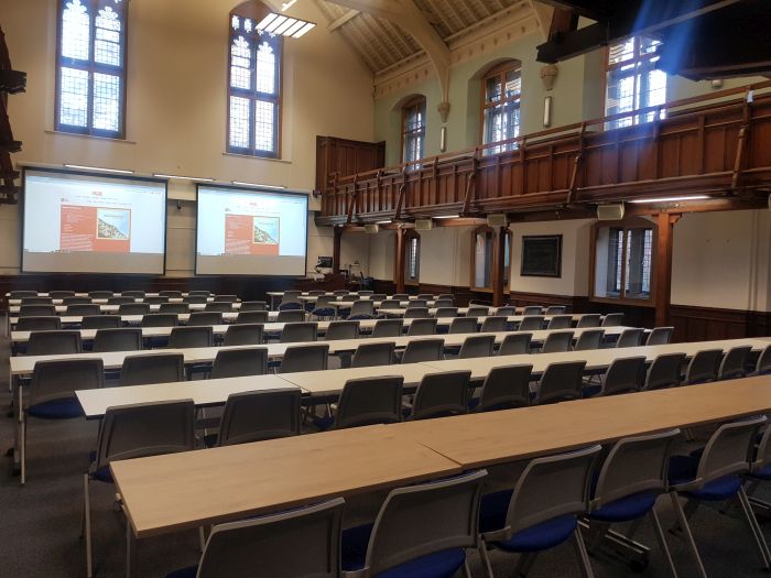 Flat floored hall with rows of tables and chairs, two screens, visualiser, lectern, and PC. 