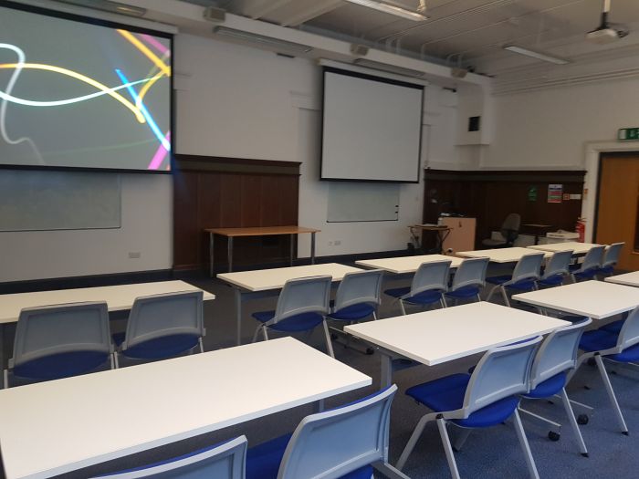 Flat floored teaching room with rows of tables and chairs, two screens, projector, and whiteboards.