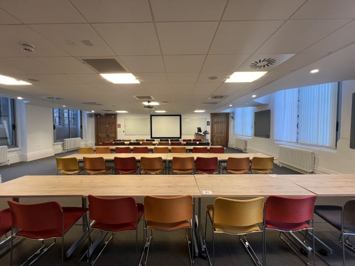 Flat floored teaching room with rows of tables and chairs, large screen, whiteboards, glassboards, projector, lectern, and PC.