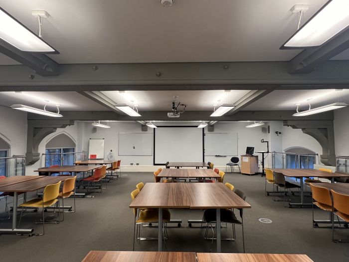 Flat floored teaching room with tables and chairs in groups, wall-mounted whiteboards, moveable whiteboard, large screen, projector, visualiser, lectern, lecturer's chair, and PC.