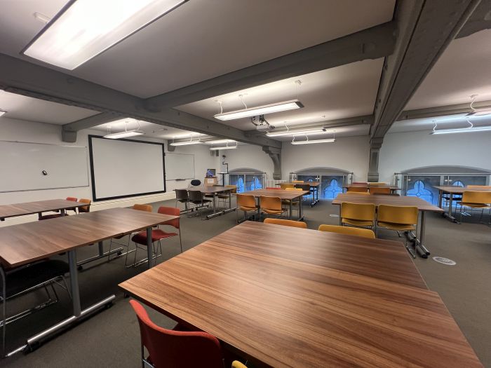 Flat floored teaching room with tables and chairs in groups, whiteboards, large screen, visualiser, lectern, and PC.