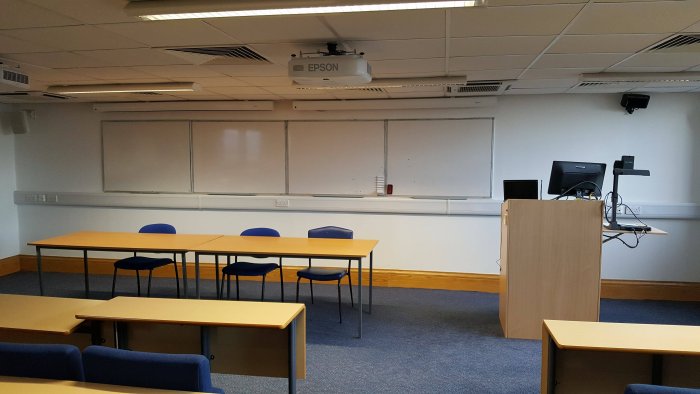 Raked lecture theatre with fixed seating, projector, whiteboards, visualiser, and PC