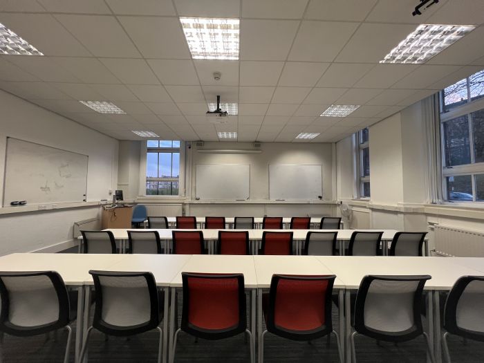 Flat floored teaching room with rows of tables and chairs, whiteboards, lectern, projector, lecturer's chair, and PC.