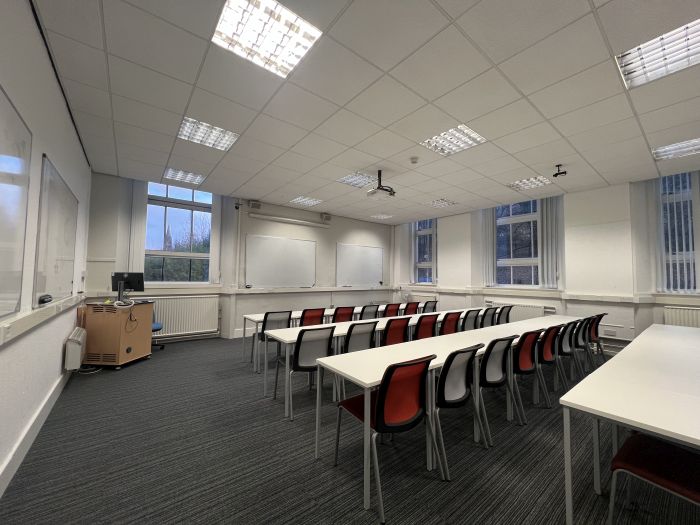 Flat floored teaching room with rows of tables and chairs, whiteboards, lectern, projector, lecturer's chair, and PC.