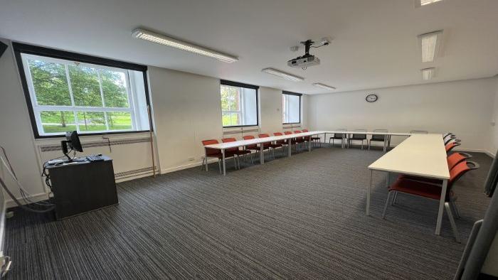 Flat floored teaching room with tables and chairs in a horseshoe formation, projector, and PC.