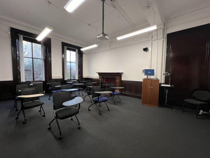 Flat floored teaching room with rows of tablet chairs, projector, lecturer's chair, visualiser, lectern, and PC.
