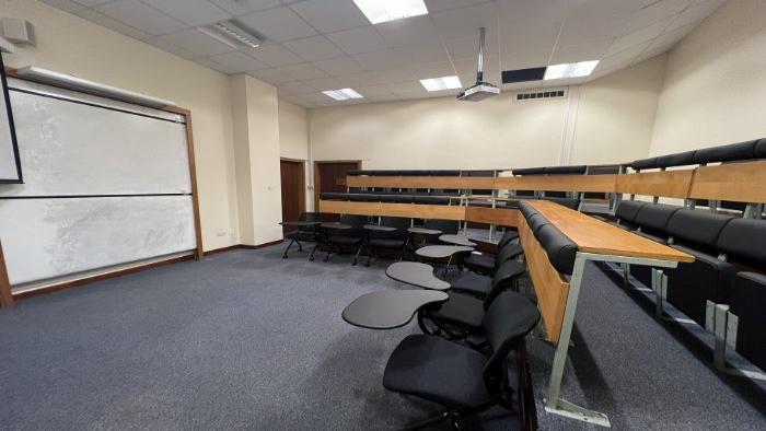 Lecture theatre with raked seating in an L-shape behind a single row of tablet chairs, whiteboard, and projector.