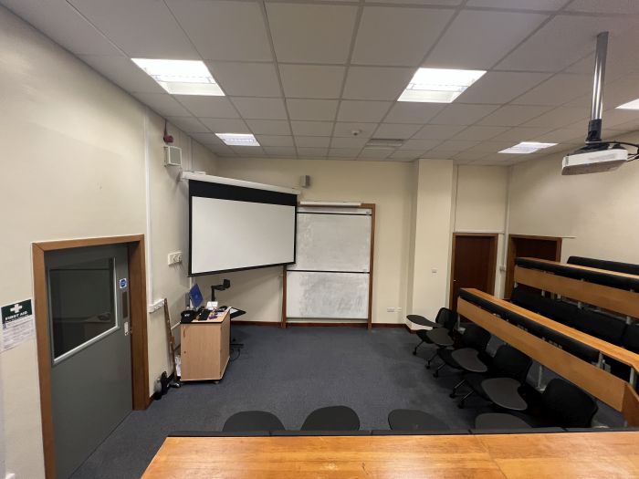 Lecture theatre with raked seating in an L-shape behind a single row of tablet chairs, whiteboard, projector, large screen, visualiser, lectern, and PC.