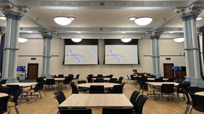 Flat floored hall with tables and chairs in groups, large screens, video monitors, lectern, and PC..