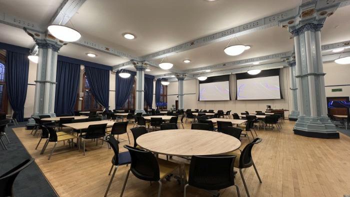Flat floored hall with tables and chairs in groups, large screens, video monitors, lectern, and PC.
