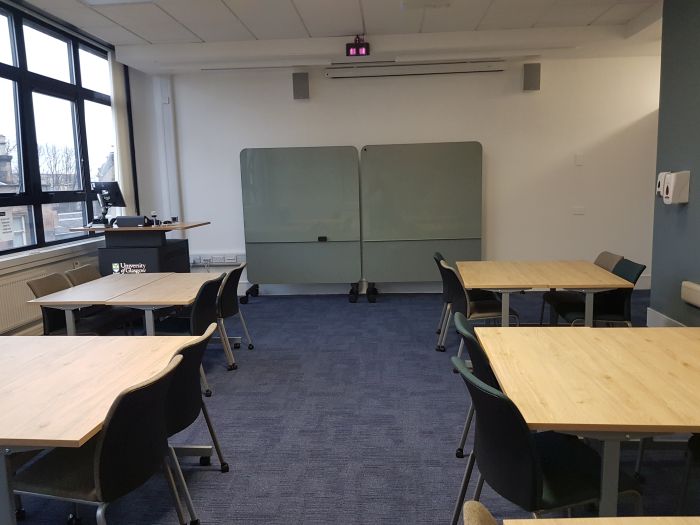 Flat floored teaching room with tables and chairs, projector, screen, PC, lectern, and moveable glassboards.