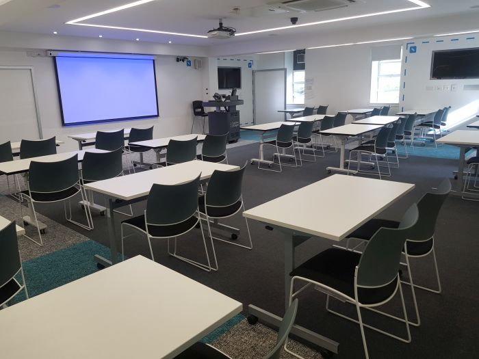 Flat floored teaching room with rows of tables and chairs, writing walls, video monitors, projector, large screen, PC, and lectern.