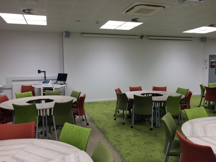 Flat floored teaching room with groups of round tables and chairs, screens, visulaiser, and PC