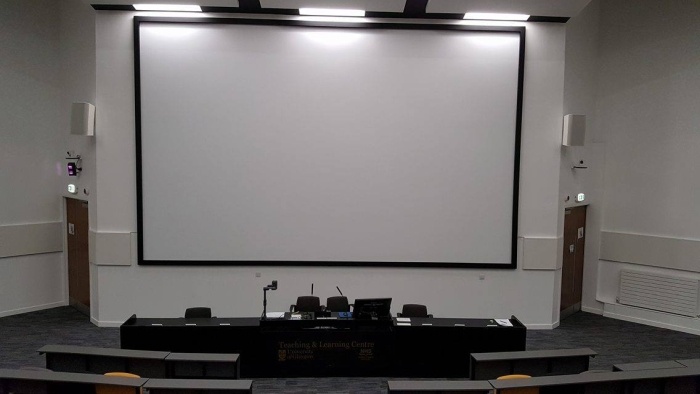 Raked lecture theatre with fixed seating, screen, visualiser, and PC