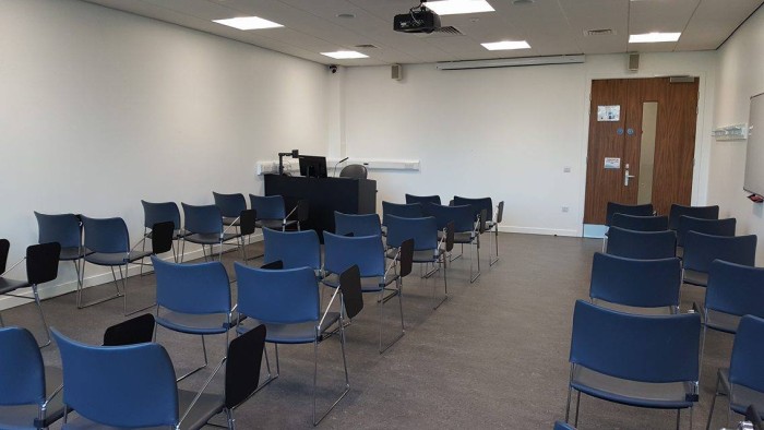 Flat floored teaching room with rows of tablet chairs, projector, screen, visualiser, and PC