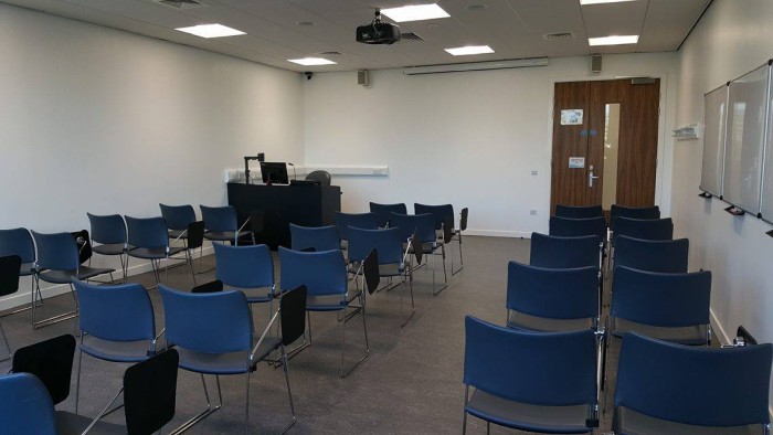 Flat floored teaching room with rows of tablet chairs, projector, screen, visualiser, and PC