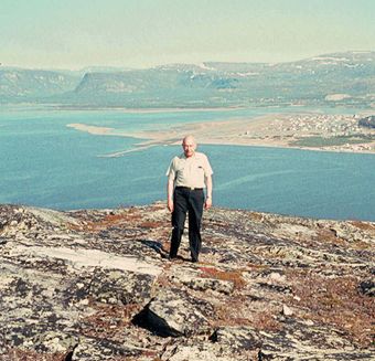 old-school photograph of an older man looking at the camera and standing on a cliff, with the sea and rocky horizon behind him