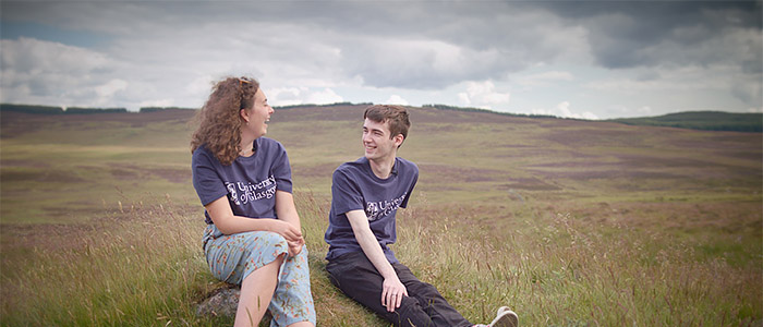 A female and male student wearing University of Glasgow t-shirts sitting in a field with hills behind them