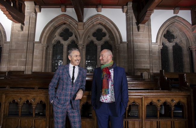 A photo of the actor Alan Cumming and theatre director John Tiffany leaning on a wooden bench in the University's Bute Hall 