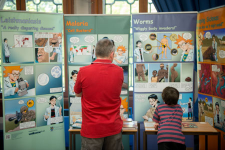 Photograph of a man and child looking at large posters which show cartoons about different diseases such as malaria. 