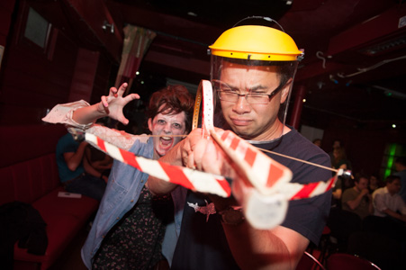 Photograph showing a man with a crossbow and a hard hat hunting zombies, in the background a zombie actor as jumped up behind him. 