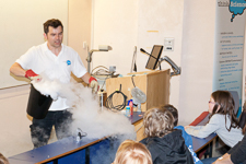 Photograph of a man demonstrating liquid nitrogen usage. There is a lot of white smoke with several children observing. 