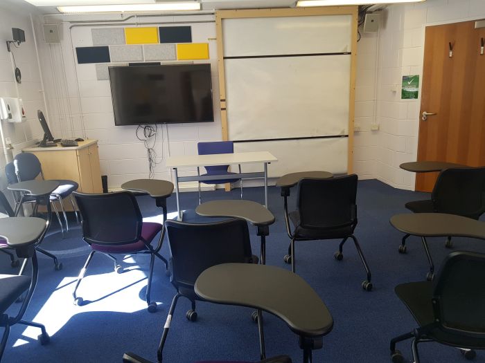 Flat floored teaching room with tablet chairs, whiteboard, large monitor, lecturer's table and chair, and PC.