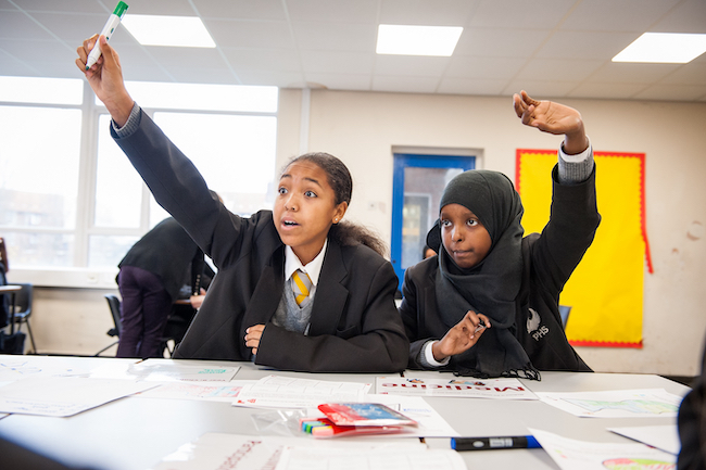 Two high school pupils raise their hands to answer a question in class. Photo courtesy of IntoUniversity