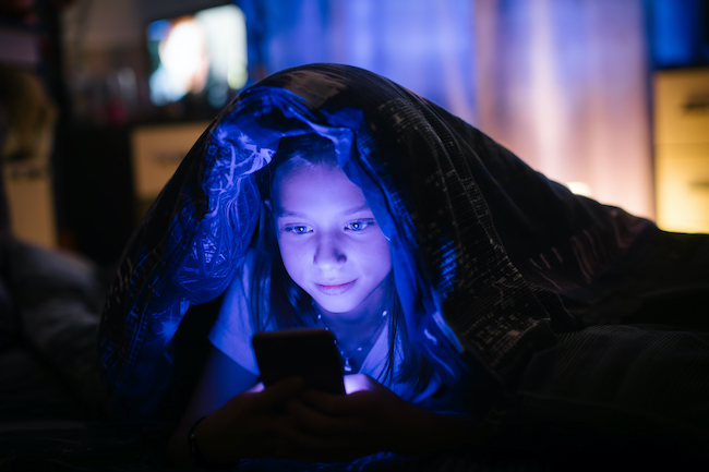 A picture of a teenager looking at her phone while under her bed covers