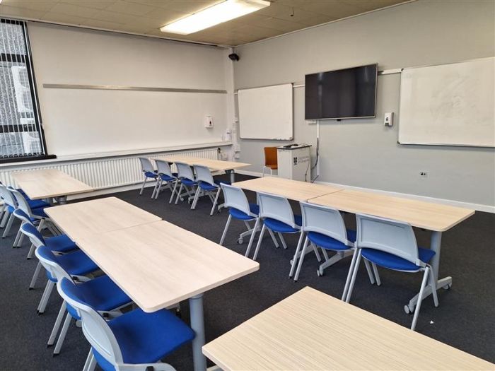 Flat floored teaching room with rows of tables and chairs, video monitor, whiteboards, lectern, and lecturer's chair..
