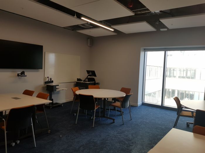 Flat floored teaching room with tables and chairs in groups, PC, lectern, large screen, and whiteboard.
