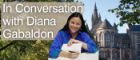 Image of University of Glasgow skyline with trees in foreground, overlaid with image of dark haired white woman writing on paper and wearing a blue shirt. Text overlay reads 'In conversation with Diana Gabaldon'