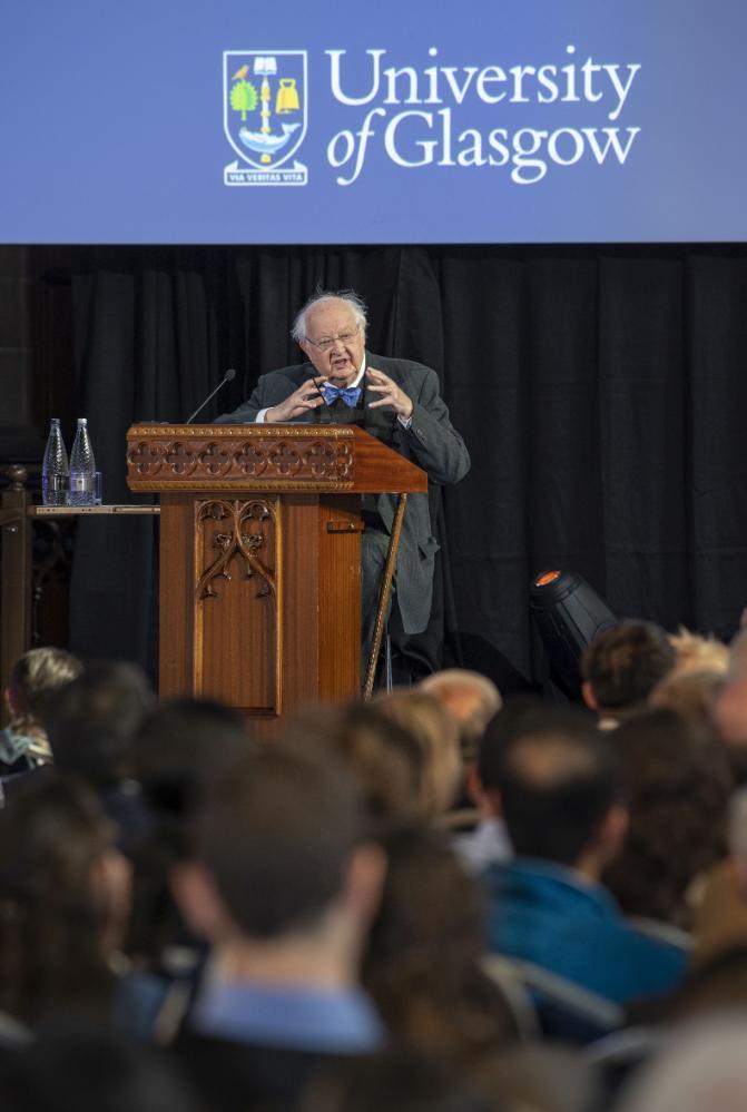Angus Deaton standing at a podium talking to a large crowd in front with the Univeristy of Glasgow logo above him. Source: Charlotte Morris, External Relations