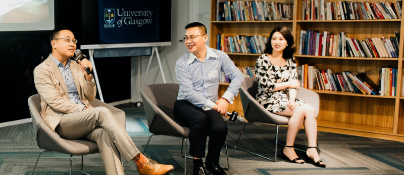 Three alumni sit on chairs before an audience, one holds a microphone while the other two smile at what he has said