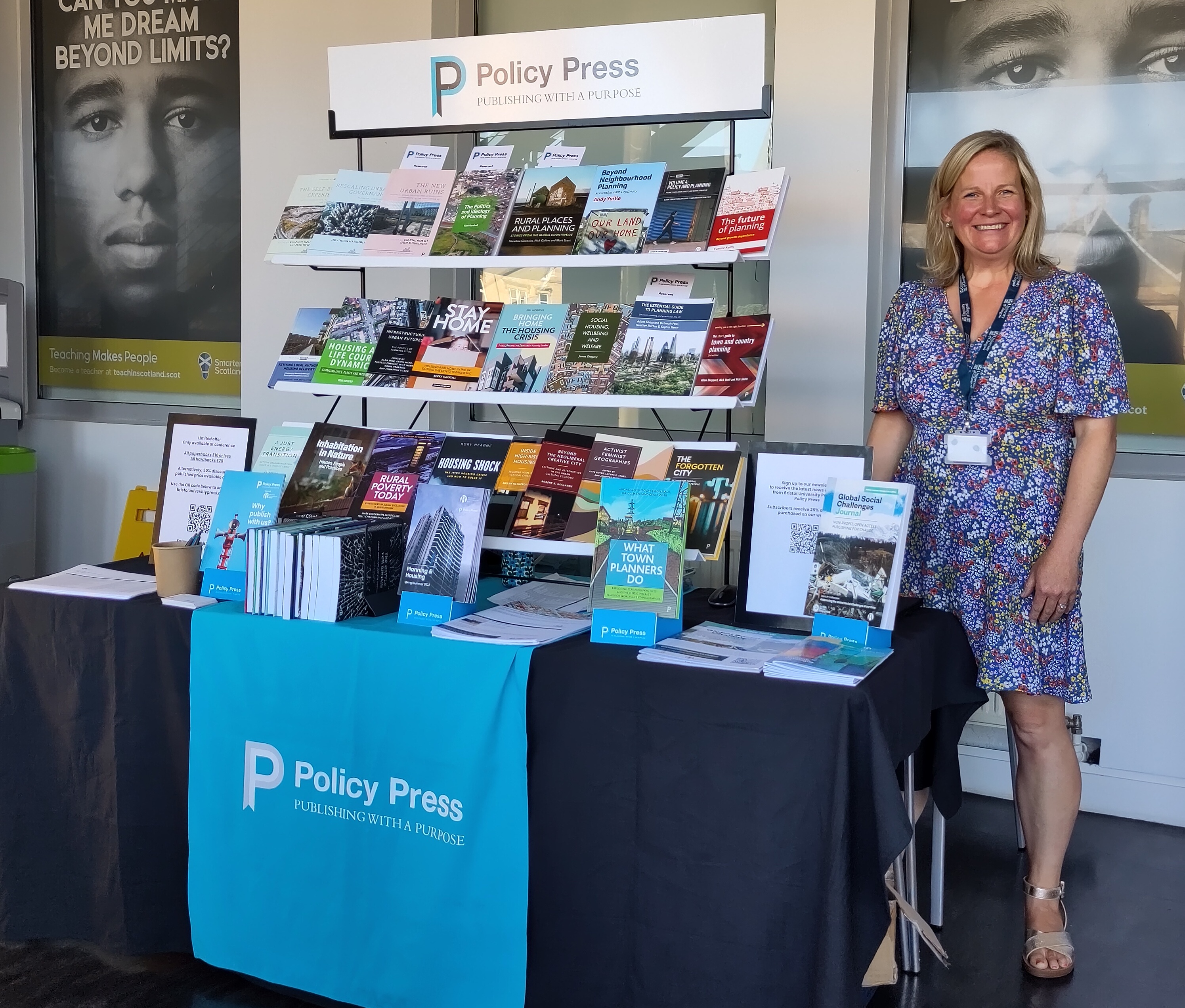 A smiling woman standing by a stall with books on display and a banner saying 'Policy Press. Publishing with a purpose'