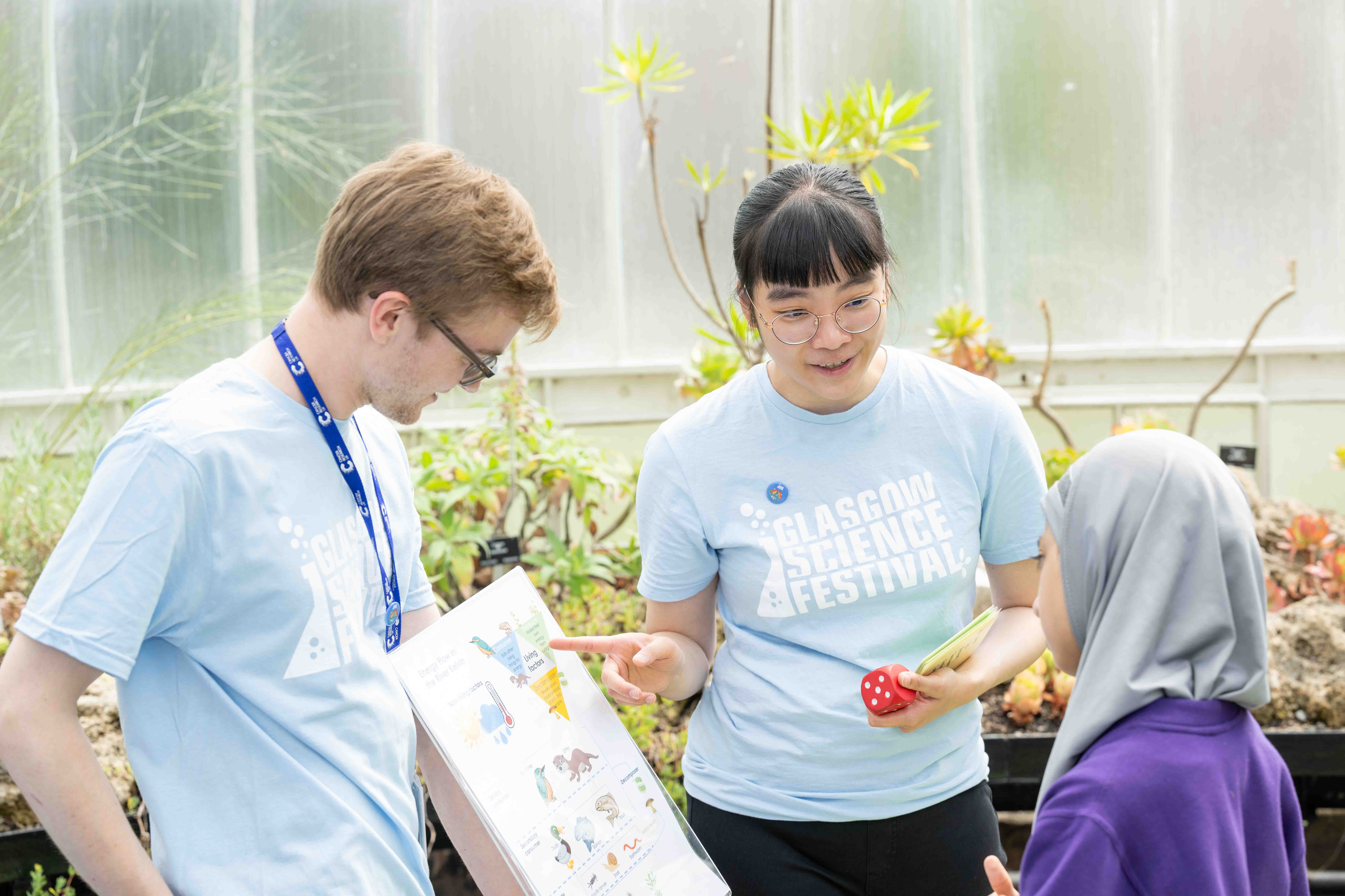 Photograph of two GSF in Action students in the Botanic Gardens with an activity participant. One person is holding a poster whilst the other is pointing to it and explaining the meaning to the participant.